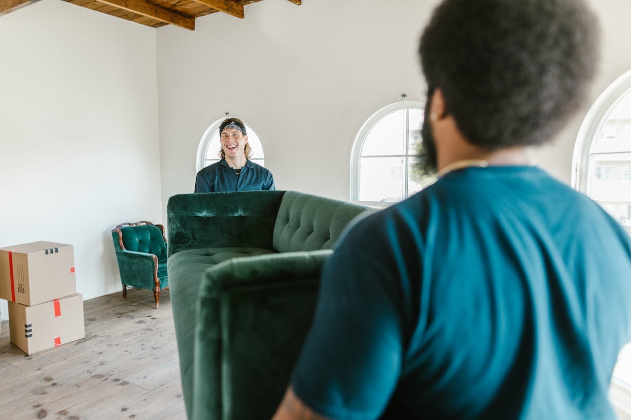 Servicios de Mudanzas en Málaga. Two movers carrying a green couch in a sunlit room with packed boxes.